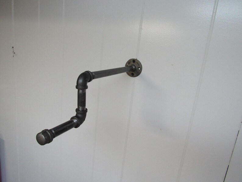 May include: Black metal pipe shelf with a flange mounted to a white wall. The shelf is bent into an L-shape.