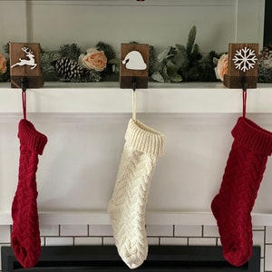 May include: Three Christmas stockings hang from a mantel, each with a different design. The stockings are red and cream-colored, with a cable knit pattern. Above them are wooden holders with festive cutouts of a reindeer, Santa hat, and snowflake.