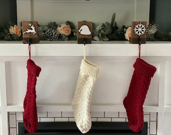 May include: Three Christmas stockings hang from a mantel, each with a different design. The stockings are red and cream-colored, with a cable knit pattern. Above them are wooden holders with festive cutouts of a reindeer, Santa hat, and snowflake.