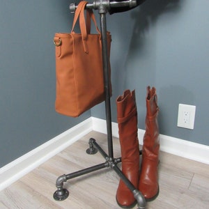 May include: A black metal coat rack with a brown leather handbag hanging from the top and two brown leather boots resting on the bottom. The rack has a four-legged base and is standing on a light wood floor.
