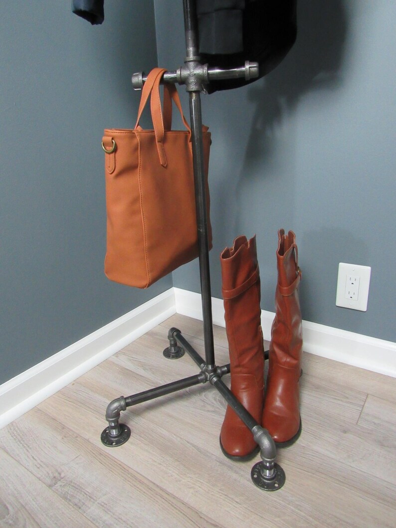May include: A black metal coat rack with a brown leather handbag hanging from the top and two brown leather boots resting on the bottom. The rack has a four-legged base and is standing on a light wood floor.