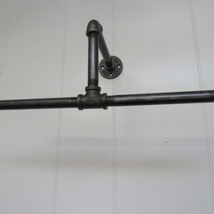 May include: Black metal pipe towel rack mounted on a white wall. The rack has a horizontal bar and a vertical support with a flange.