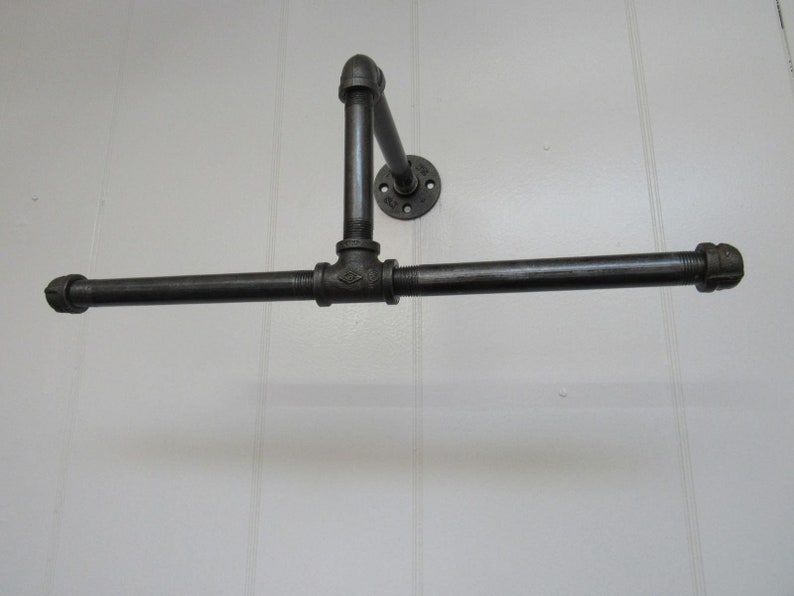 May include: Black metal pipe towel rack mounted on a white wall. The rack has a horizontal bar and a vertical support with a flange.
