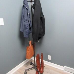 May include: A black metal coat rack with a brown leather bag hanging on the bottom and two pairs of brown boots on the floor. The rack is in a corner of a room with gray walls and light wood floors.