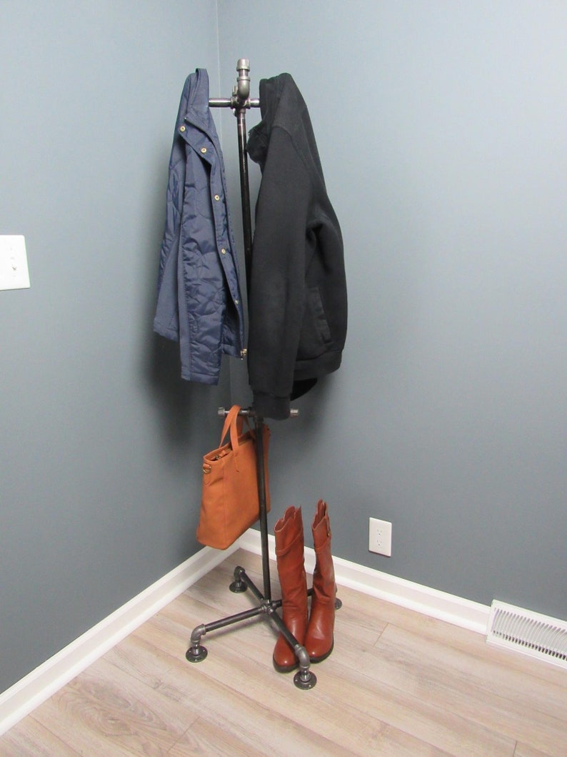 May include: A black metal coat rack with a brown leather bag hanging on the bottom and two pairs of brown boots on the floor. The rack is in a corner of a room with gray walls and light wood floors.