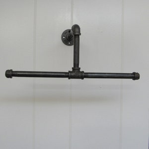 May include: A black metal pipe towel rack with a flange mounted to a white wall. The rack has a horizontal bar and a vertical bar that extends from the wall.