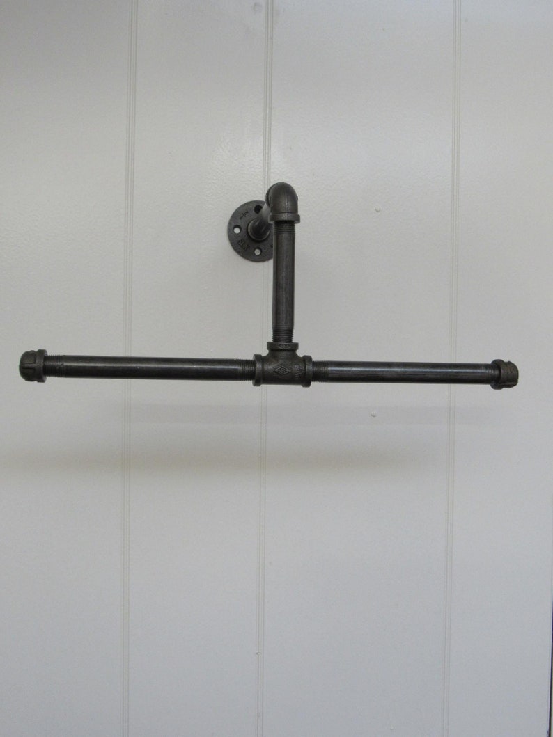 May include: A black metal pipe towel rack with a flange mounted to a white wall. The rack has a horizontal bar and a vertical bar that extends from the wall.
