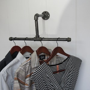 May include: A black metal pipe clothes rack mounted on a white wall with four wooden hangers holding a striped shirt, a patterned shirt, a black and white striped dress, and a dark blue blazer.