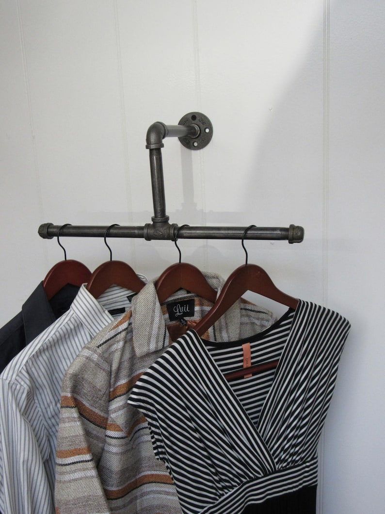 May include: A black metal pipe clothes rack mounted on a white wall with four wooden hangers holding a striped shirt, a patterned shirt, a black and white striped dress, and a dark blue blazer.