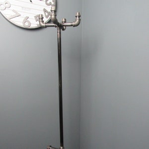 May include: A black metal coat rack with four arms extending from a central pole. The arms are made of pipe fittings and have a silver finish. The rack is standing in a corner of a room.