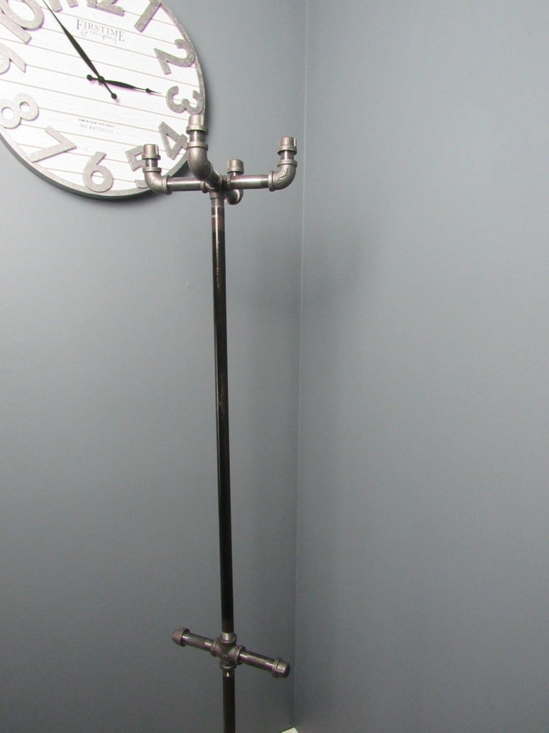 May include: A black metal coat rack with four arms extending from a central pole. The arms are made of pipe fittings and have a silver finish. The rack is standing in a corner of a room.