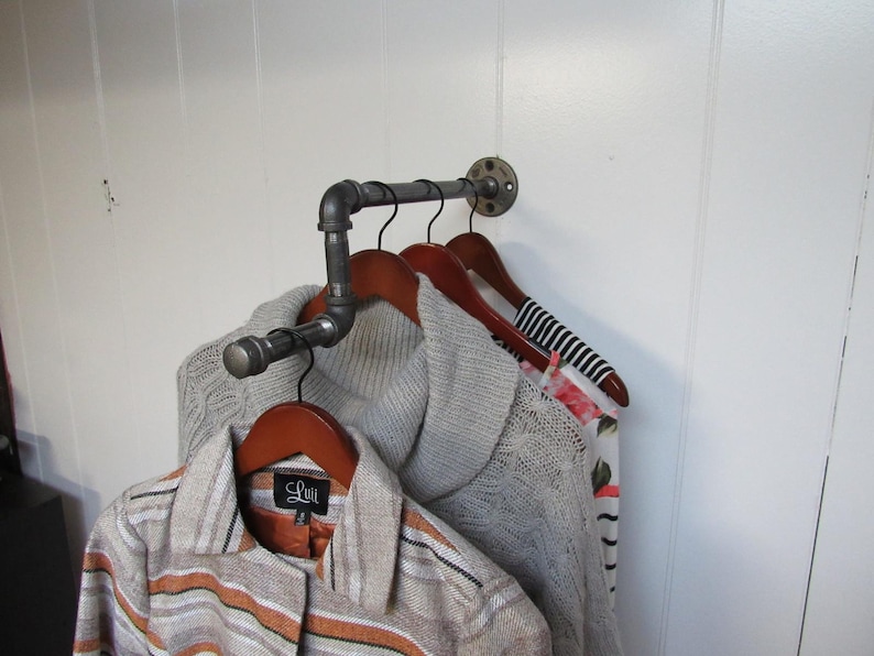 May include: A metal pipe clothes rack with four wooden hangers holding sweaters and a shirt. The rack is mounted to a white wall.