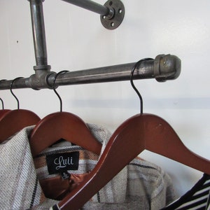 May include: A black metal pipe clothes rack with two wooden hangers and two shirts hanging on them. One shirt has a brown and white plaid pattern and a label that says "Luii". The other shirt is black and white striped.