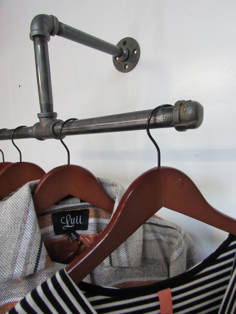 May include: A black metal pipe clothes rack with two wooden hangers and two shirts hanging on them. One shirt has a brown and white plaid pattern and a label that says "Luii". The other shirt is black and white striped.