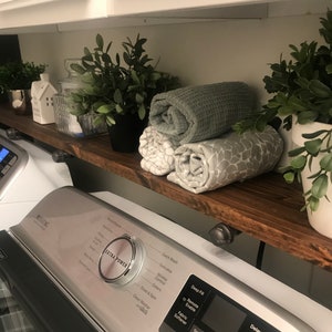 May include: A white washing machine with a silver control panel and a wooden shelf above it. The shelf is decorated with potted plants, rolled towels, and a small white house figurine. The washing machine has a variety of settings, including "Extra Power", "Quick Wash", "Delicates", "Wrinkle Control", "Colors", "Drain & Spin", "Deep Fill", "Remote Enable", "Fabric Softener", "Spin", "Temp", "Soil", "Delay Start", "Extra Rinse", and "Cycle Signal".