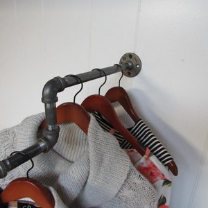 May include: A black metal pipe clothes rack with three wooden hangers and clothing hanging on them. The rack is mounted to a white wall.