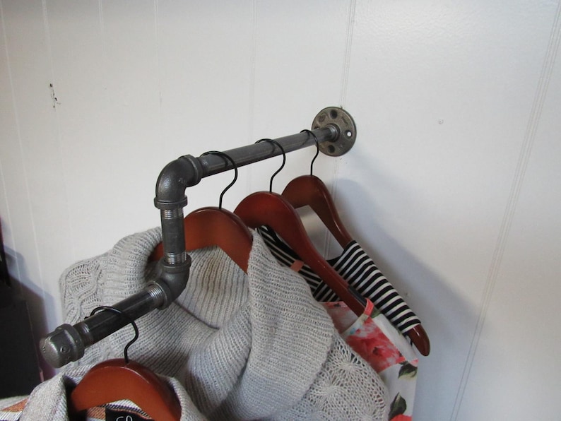 May include: A black metal pipe clothes rack with three wooden hangers and clothing hanging on them. The rack is mounted to a white wall.