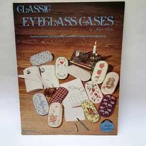May include: A book cover titled "CLASSIC EYEGLASS CASES" by Joyce Seebo. The cover features various cross-stitch eyeglass case designs, including floral, heart, and owl motifs. The book is on a wooden surface with other craft supplies.