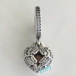 May include: A silver charm with a heart-shaped pendant featuring wing-like details and embedded clear stones. The charm has a ring with clear stones around it. The pendant is approximately 0.75 inches wide.
