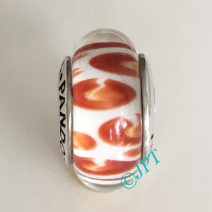 May include: A clear glass bead with a white and orange pattern. The bead has a cylindrical shape with silver-colored metal accents. The word "PANDORA" is visible on one side of the bead.