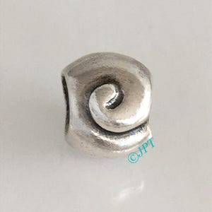 May include: A silver-toned charm with a spiral design. The charm has a rounded shape with a swirl detail in the center. The surface has a slightly textured appearance.