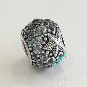 May include: A spherical silver charm bead featuring a textured starfish relief and numerous embedded round, translucent blue and clear glass stones. The surface is detailed with small silver spheres between the stones. The text JPT is visible.