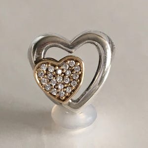 May include: A silver-toned heart-shaped pendant with a smaller gold-toned heart at its center. The smaller heart is encrusted with numerous small, clear gemstones. The pendant is displayed on a white surface.