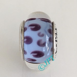 May include: A glass bead with a light blue center and dark purple oval shapes. The bead has silver-colored metal accents on each end. The text "PA E" is visible on the metal. The bead is a decorative jewelry item.