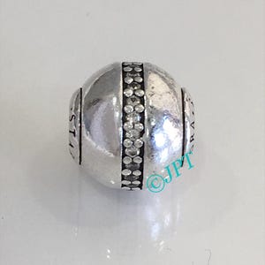 May include: A silver bead with a band of clear, sparkling stones around the center. The bead has a rounded shape and features engraved text on each side. A jewelry item.