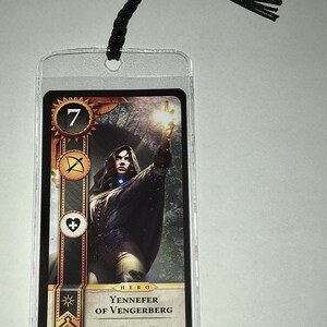 Peut inclure: Une carte de jeu avec une illustration de Yennefer de Vengerberg, avec le chiffre 7 et des symboles. La carte est dans une pochette en plastique transparent et est attach&eacute;e &agrave; un cordon noir en haut.