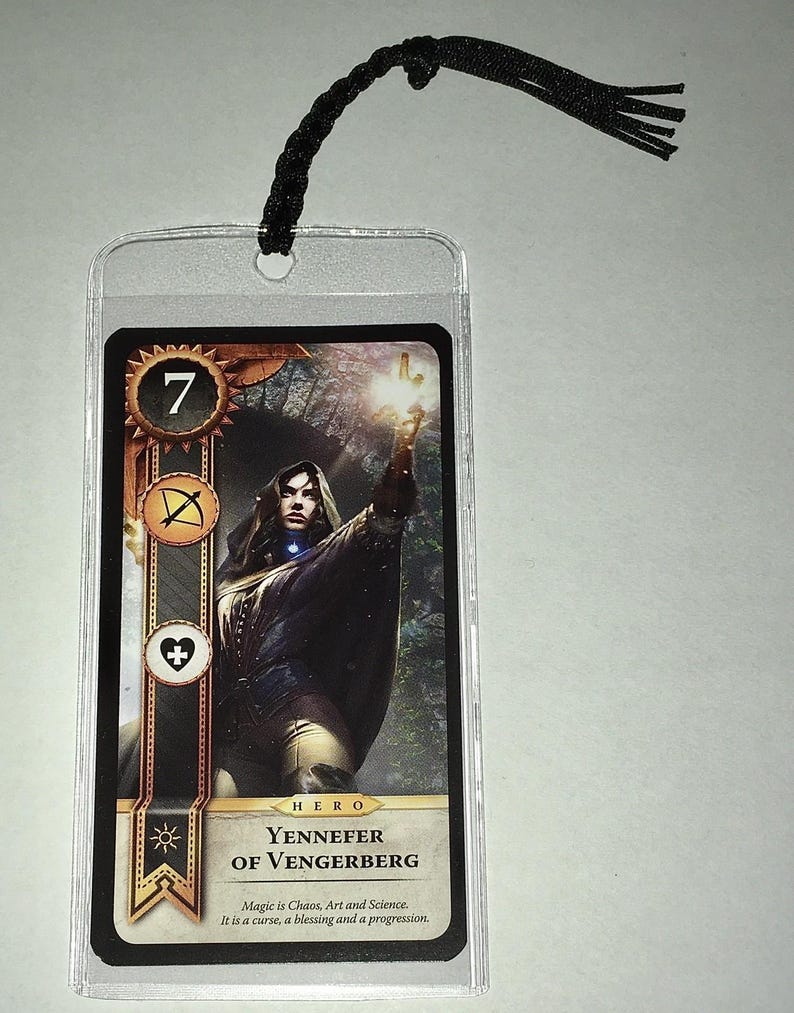 Peut inclure: Une carte de jeu avec une illustration de Yennefer de Vengerberg, avec le chiffre 7 et des symboles. La carte est dans une pochette en plastique transparent et est attach&eacute;e &agrave; un cordon noir en haut.