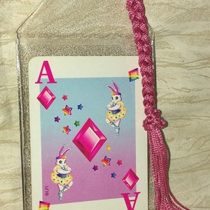 May include: A playing card showing the Ace of Diamonds, with a playful design of dancing rabbits. The card is in a clear plastic sleeve, with a pink braided cord and tassel, designed to be used as a bookmark.