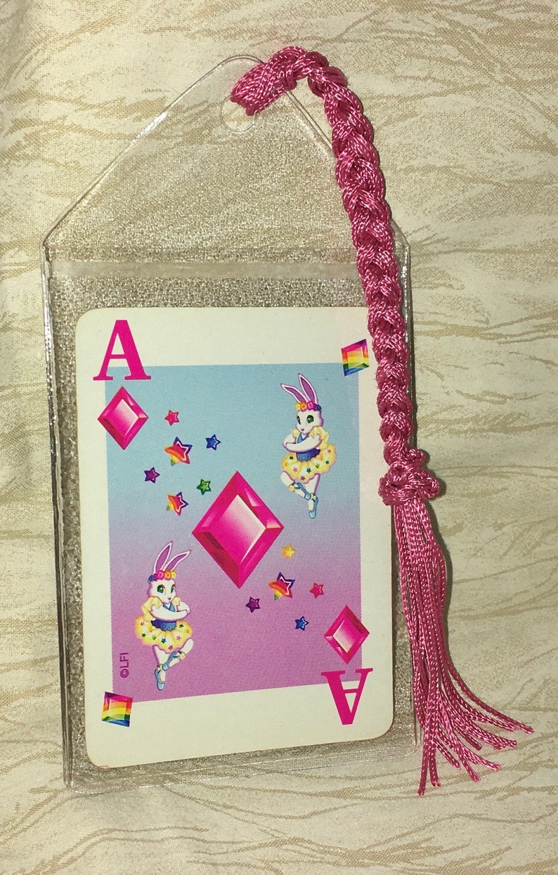 May include: A playing card showing the Ace of Diamonds, with a playful design of dancing rabbits. The card is in a clear plastic sleeve, with a pink braided cord and tassel, designed to be used as a bookmark.