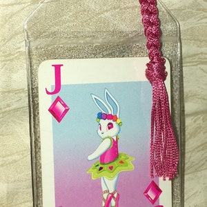 May include: A clear plastic bookmark with a playing card design. The card is a Jack of Diamonds, featuring a cartoon rabbit ballerina. A pink braided tassel is attached. The rabbit wears a tutu and flower crown.