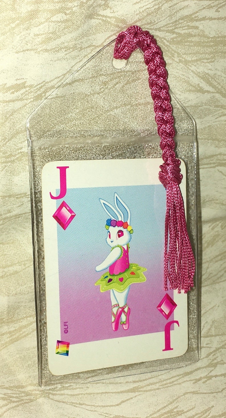 May include: A clear plastic bookmark with a playing card design. The card is a Jack of Diamonds, featuring a cartoon rabbit ballerina. A pink braided tassel is attached. The rabbit wears a tutu and flower crown.