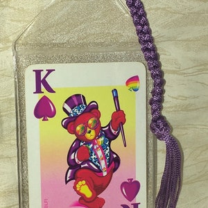 May include: A playing card featuring a cartoon bear in a purple suit and top hat, holding a wand. The card is the King of Spades, in a clear plastic sleeve with a purple tassel.