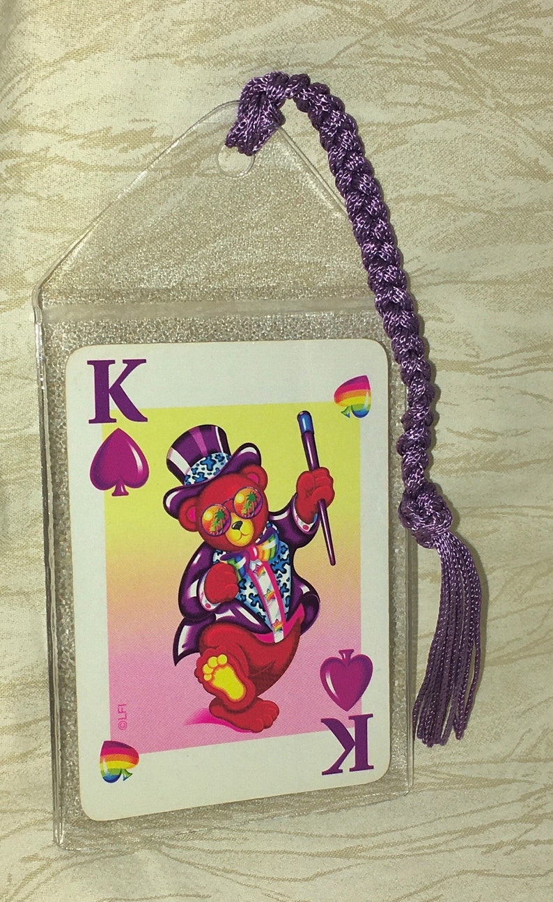 May include: A playing card featuring a cartoon bear in a purple suit and top hat, holding a wand. The card is the King of Spades, in a clear plastic sleeve with a purple tassel.