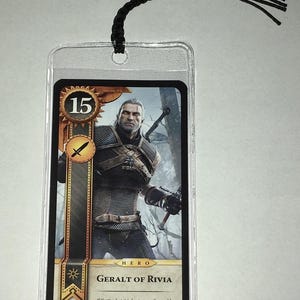 Peut inclure: Marque-page en plastique transparent avec une carte repr&eacute;sentant Geralt de Riv, personnage de la s&eacute;rie The Witcher. La carte a un bord dor&eacute;, le chiffre 15 et le texte "GERALT OF RIVIA". Une corde noire est attach&eacute;e en haut.