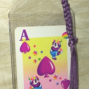 May include: A clear plastic bookmark with a playing card design. The card features a purple Ace of Spades and cartoon pandas with rainbow accents. A braided purple cord and tassel are attached.