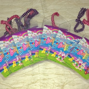 May include: A selection of Lisa Frank bookmarks in clear plastic sleeves. Each bookmark displays a vibrant design with rainbows, unicorns, and puppies. The bookmarks have braided, glittery cords in shades of purple and pink.