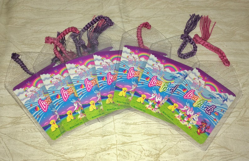 May include: A selection of Lisa Frank bookmarks in clear plastic sleeves. Each bookmark displays a vibrant design with rainbows, unicorns, and puppies. The bookmarks have braided, glittery cords in shades of purple and pink.