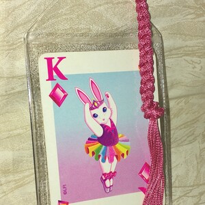 May include: A bookmark featuring a playing card with a cartoon bunny ballerina in a tutu. The card is encased in a clear plastic sleeve and has a pink braided tassel attached. The card is the King of Diamonds.