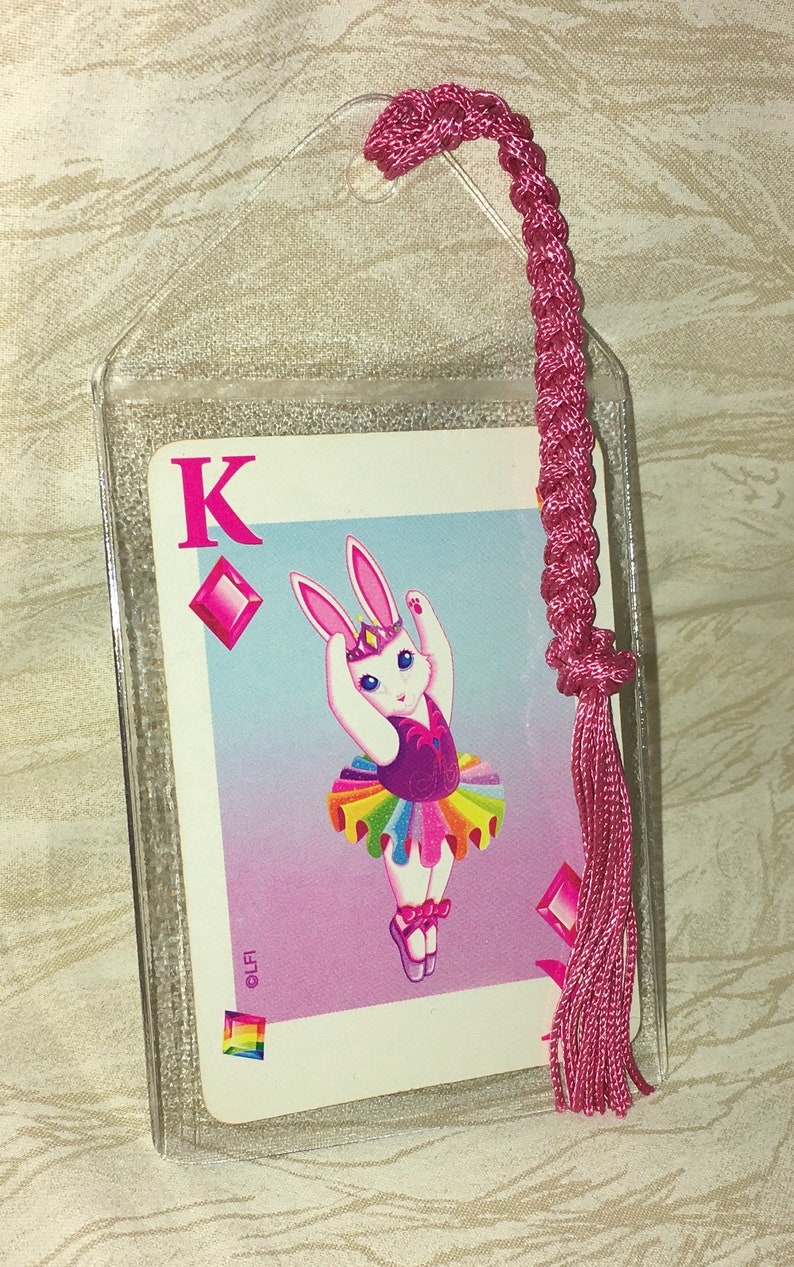 May include: A bookmark featuring a playing card with a cartoon bunny ballerina in a tutu. The card is encased in a clear plastic sleeve and has a pink braided tassel attached. The card is the King of Diamonds.