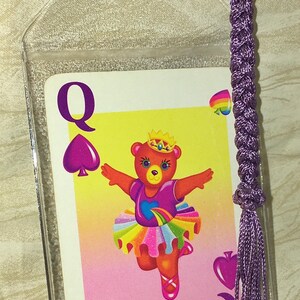 May include: A Queen of Spades playing card with a dancing bear in a tutu, inside a clear plastic sleeve. The card has a purple spade symbol and the letter "Q". A braided purple cord with a tassel is attached to the sleeve.