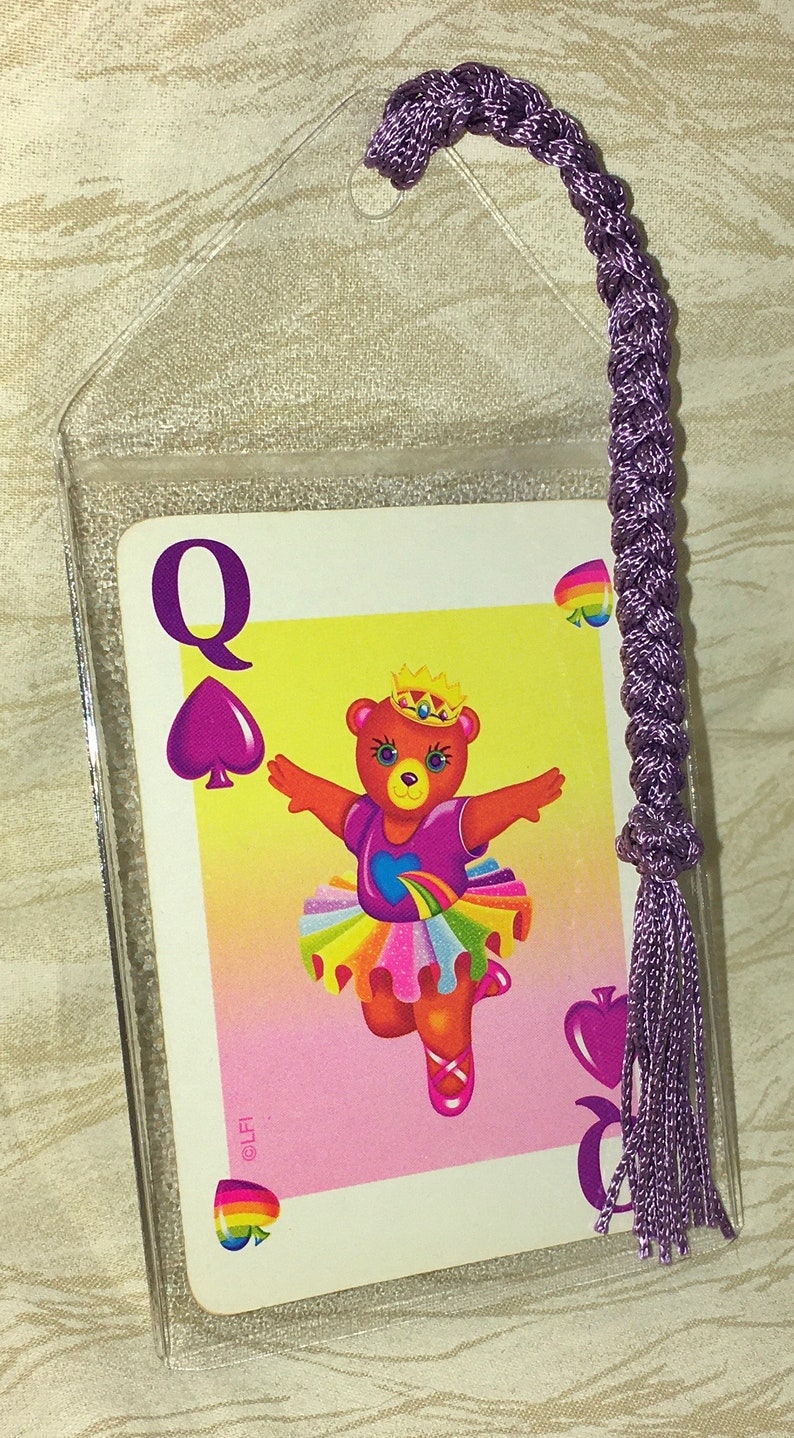 May include: A Queen of Spades playing card with a dancing bear in a tutu, inside a clear plastic sleeve. The card has a purple spade symbol and the letter "Q". A braided purple cord with a tassel is attached to the sleeve.
