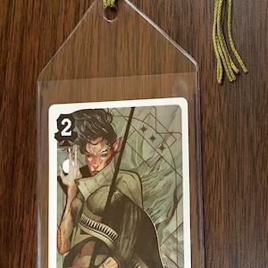 May include: A clear plastic bookmark with a green tassel. The bookmark has a slot to hold a card. The card shows a fantasy illustration of a person with pointed ears and a staff. The card has the number "2" on it.
