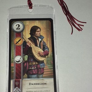 Peut inclure: Un marque-page avec une carte repr&eacute;sentant un homme jouant du luth, intitul&eacute; "DANDELION". La carte est prot&eacute;g&eacute;e dans une pochette en plastique transparent et poss&egrave;de un pompon bordeaux attach&eacute; en haut. La carte a une bande rouge et blanche sur le c&ocirc;t&eacute; gauche.
