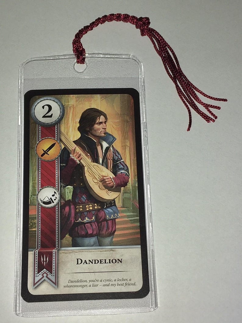 Peut inclure: Un marque-page avec une carte repr&eacute;sentant un homme jouant du luth, intitul&eacute; "DANDELION". La carte est prot&eacute;g&eacute;e dans une pochette en plastique transparent et poss&egrave;de un pompon bordeaux attach&eacute; en haut. La carte a une bande rouge et blanche sur le c&ocirc;t&eacute; gauche.