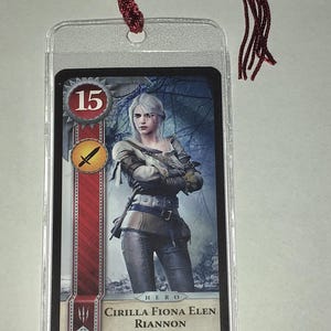 Peut inclure: Une carte &agrave; collectionner avec une illustration de personnage, ins&eacute;r&eacute;e dans une pochette en plastique transparent et une corde tress&eacute;e rouge. La carte affiche le num&eacute;ro 15, une ic&ocirc;ne d'&eacute;p&eacute;e et le nom "CIRILLA FIONA ELEN RIANNON".