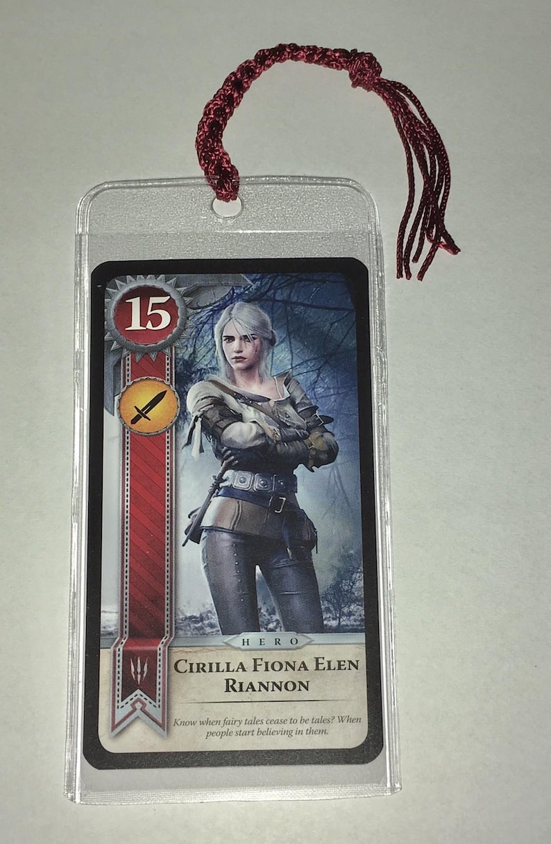 Peut inclure: Une carte &agrave; collectionner avec une illustration de personnage, ins&eacute;r&eacute;e dans une pochette en plastique transparent et une corde tress&eacute;e rouge. La carte affiche le num&eacute;ro 15, une ic&ocirc;ne d'&eacute;p&eacute;e et le nom "CIRILLA FIONA ELEN RIANNON".